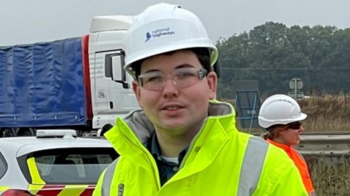 James Owen Thomas wearing a helmet and a hi-vis jacket on a section of closed road, with another person and two vehicles behind him