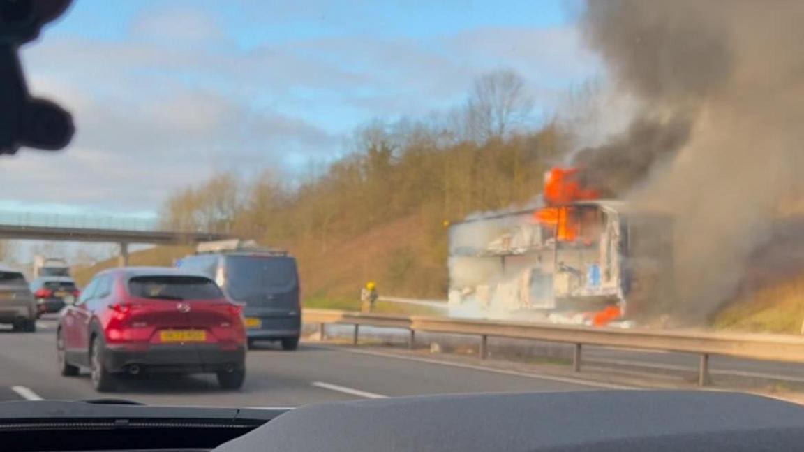 An image of a lorry on fire on the motorway, as taken from another vehicle. The lorry is already significantly burnt in this picture, with flames and smoke seen rising from the top. A dashboard is seen at the bottom of the picture, and cars can be seen travelling on the opposite side of the motorway to the lorry.