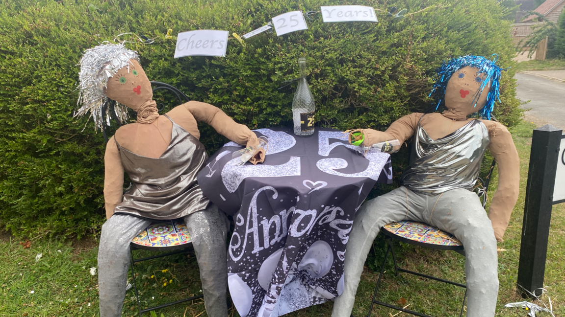 Two scarecrows, dressed as women in sparkly silver tops, are sat at a table with a banner that says 25 anniversary. One scarecrow has silver tinsel type hair and the other metallic blue. They are next to a street sign. 