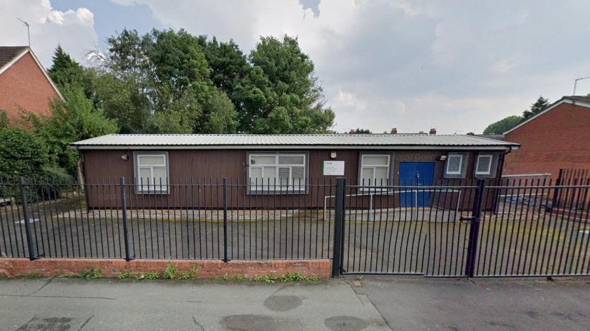 A single-storey building with a black fence separating an exercise yard from the pavement. The building has a set of blue double doors and a series of windows and wooden cladding.
