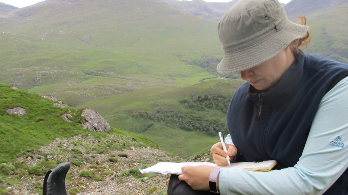 Claire Dashwood wearing a brown bucket style hat, navy gilet and baby blue long-sleeved top, writing in a notepad. She is sitting on a cliff and there are rolling green and rocky hills behind her.