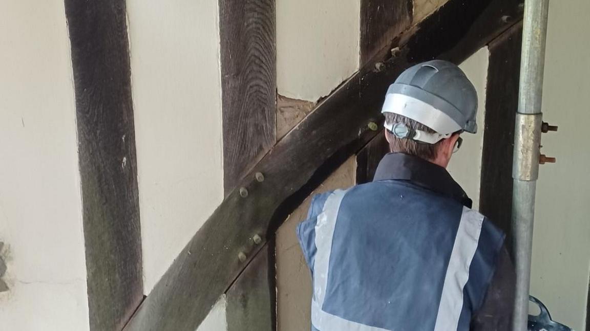A man in a navy work jacket with fluorescent white strips on the black, wearing a hard hat. He is crouched with his back to the camera and appears to be working on a cream and timber-framed wall.