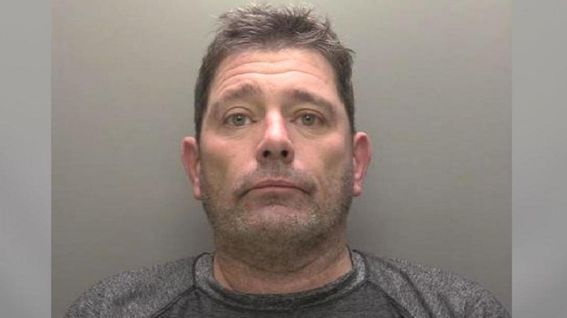 A police custody picture showing the head and shoulders of a man with short brown hair and stubble wearing a grey T-shirt against a grey backdrop.