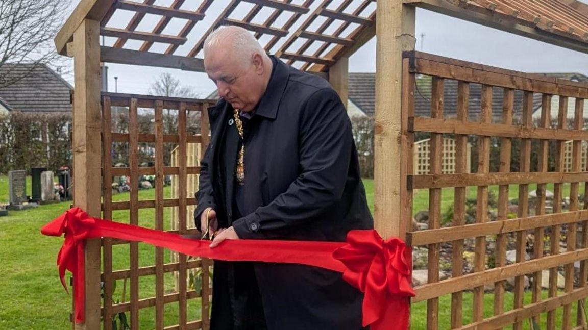 Lord Mayor of Stoke-on-Trent, Steve Watkins is stood under a wooden trellis with a red ribbon attached. He is holding a pair of scissors, and is cutting the ribbon.