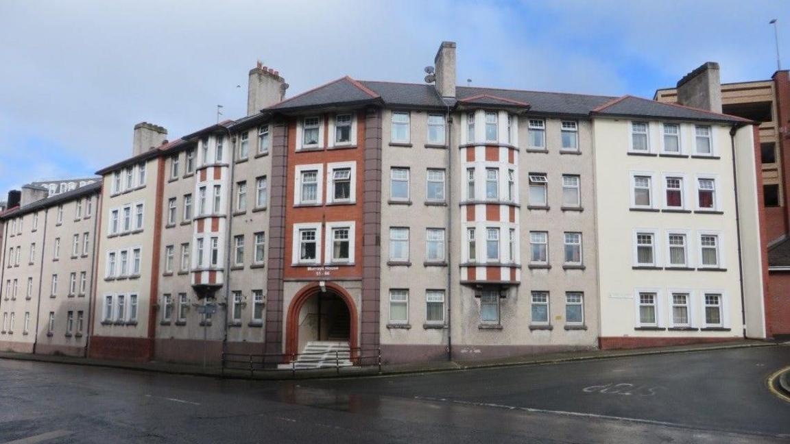 The exterior of the Lord Street flats, an aged block of flats, which has an archway entrance with steps leading into it on the corner of two roads.