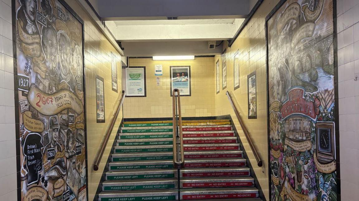 Two colourful murals line the side of two wall at a train station. There are two sets of stairs, one green, the other red.