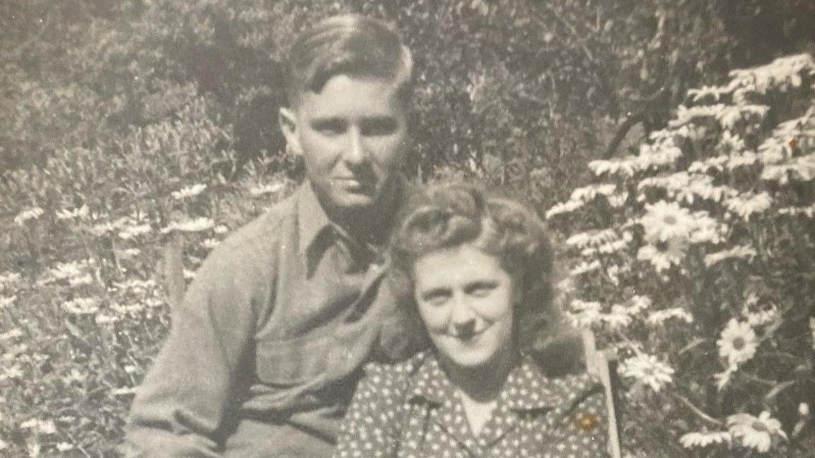 GI Mike Browning and Jackie Atwell sitting together in black and white photograph in front of flowers.