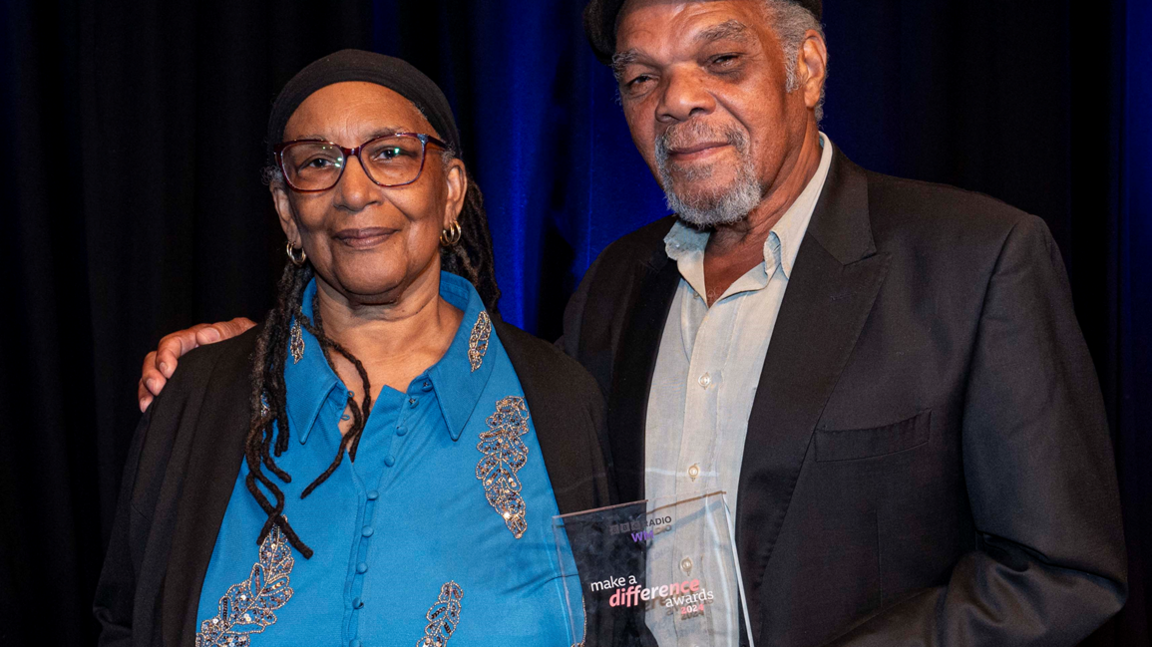 Marva is wearing a blue top and black jacket and had long hair. Lionel is in a black suit and light shirt and cap with his arm round Marva and holding their award