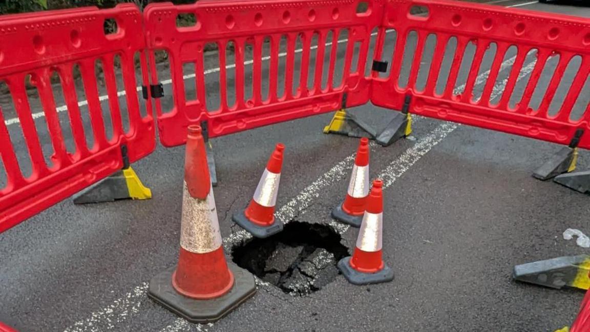 The sinkhole on the A361. It is surrounded by orange and white road cones and road barriers.