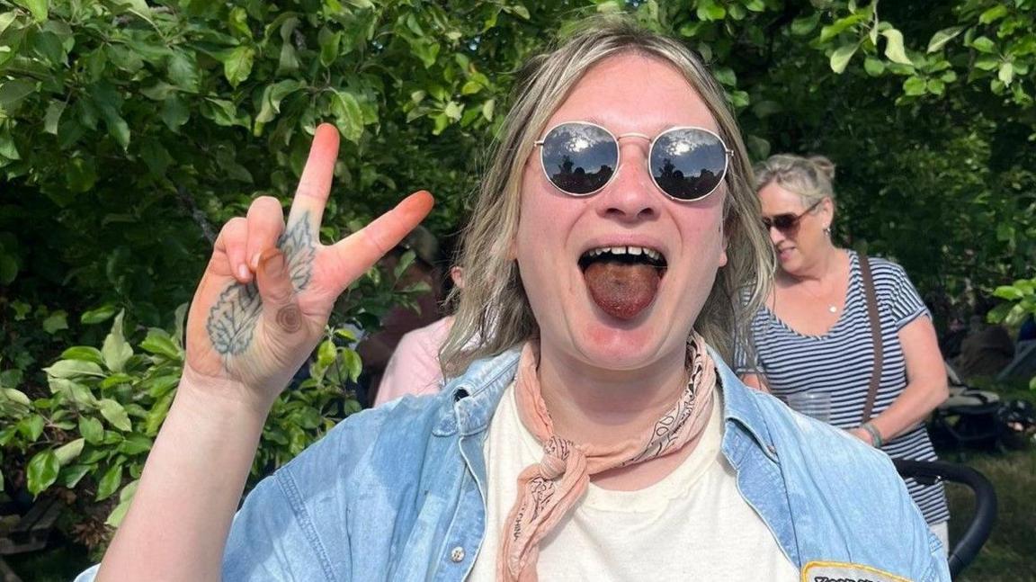 A woman holding up her right hand in a peace sign and poking out her tongue which is a green-black colour