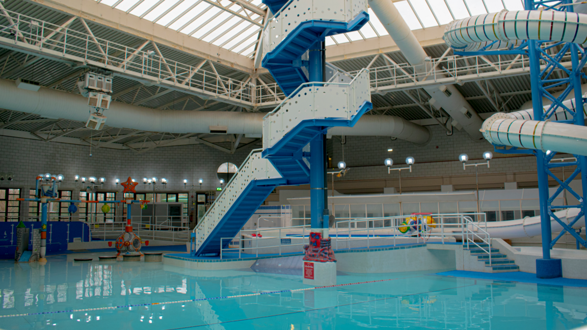 An indoor swimming pool. There is a spiral tower of steps leading to a slide. The pool is aqua blue.