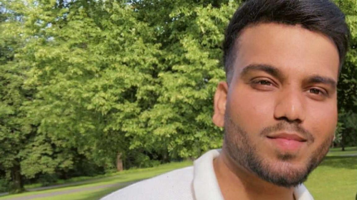  A person is standing in a grassy park with lush green trees in the background. The individual is wearing a short-sleeved white polo shirt. The lighting suggests a sunny day, and the image includes text overlay that reads “Armaan Wednesday” in white letters near the lower left side.