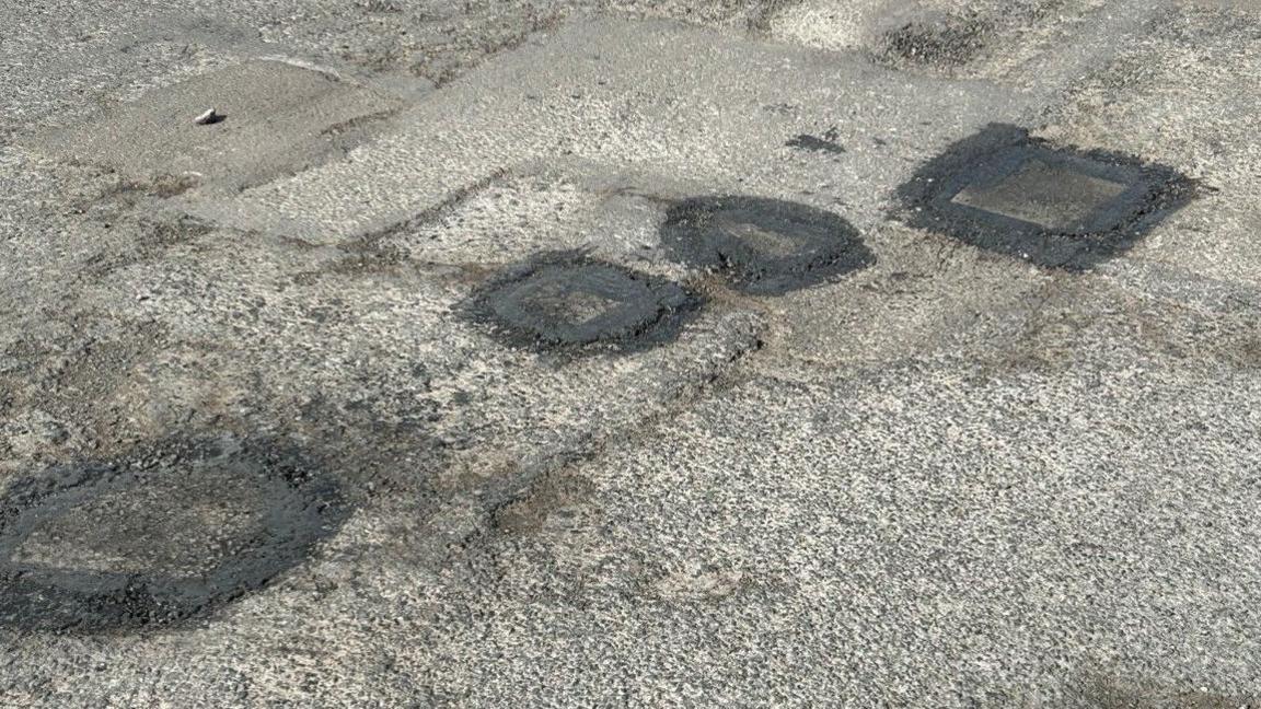 Four potholes filled in with asphalt on a road. They go from bottom left to top right on the image.