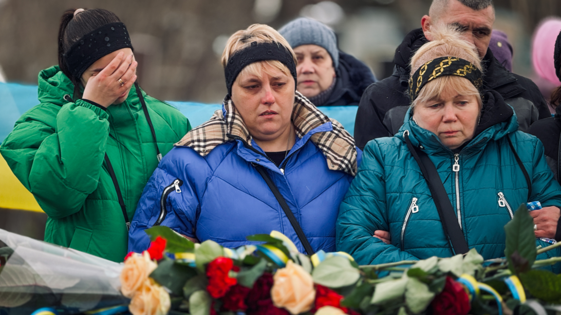 Natalya is flanked by two other women at her husband's funeral, with flowers visible in the foreground.