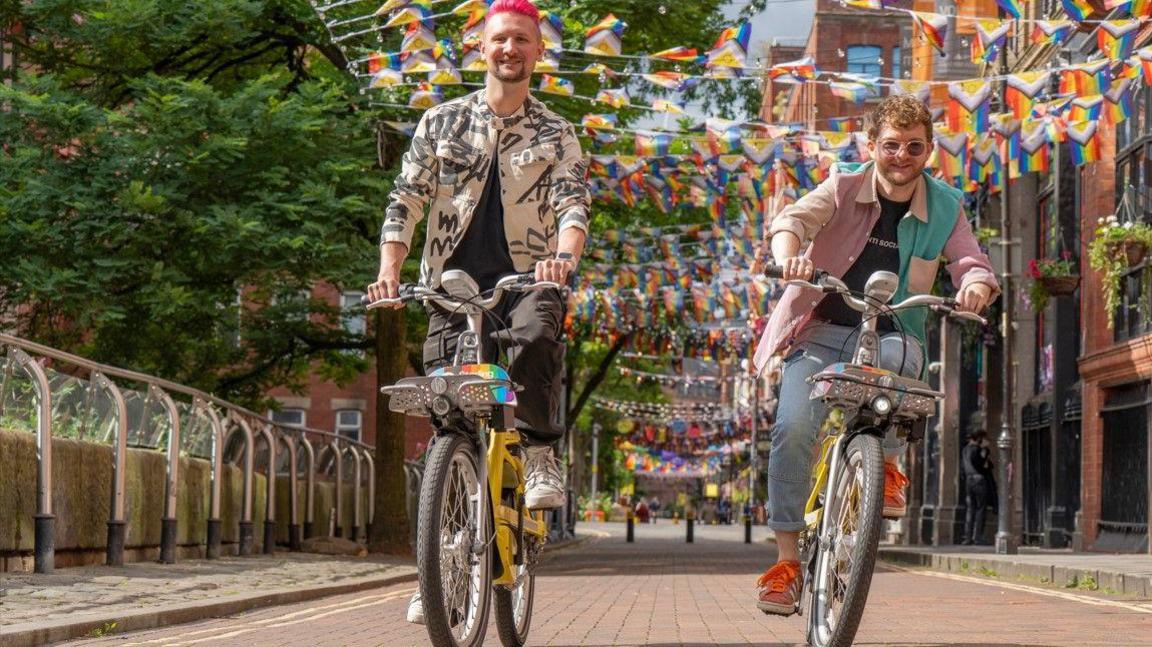 A person with pink hair and a patterned black and white jacket and black trousers and another person with light brown hair and sunglasses and wearing a pink and green shirt and blue jeans are smiling while riding Starling bikes. The bikes have Pride branding, with Pride bunting seen in the street above them.