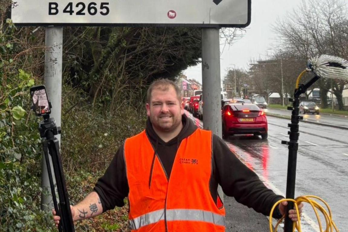 Man in front of a road sign holding tripod and window cleaning equipment. He is stood next to a busy main road.