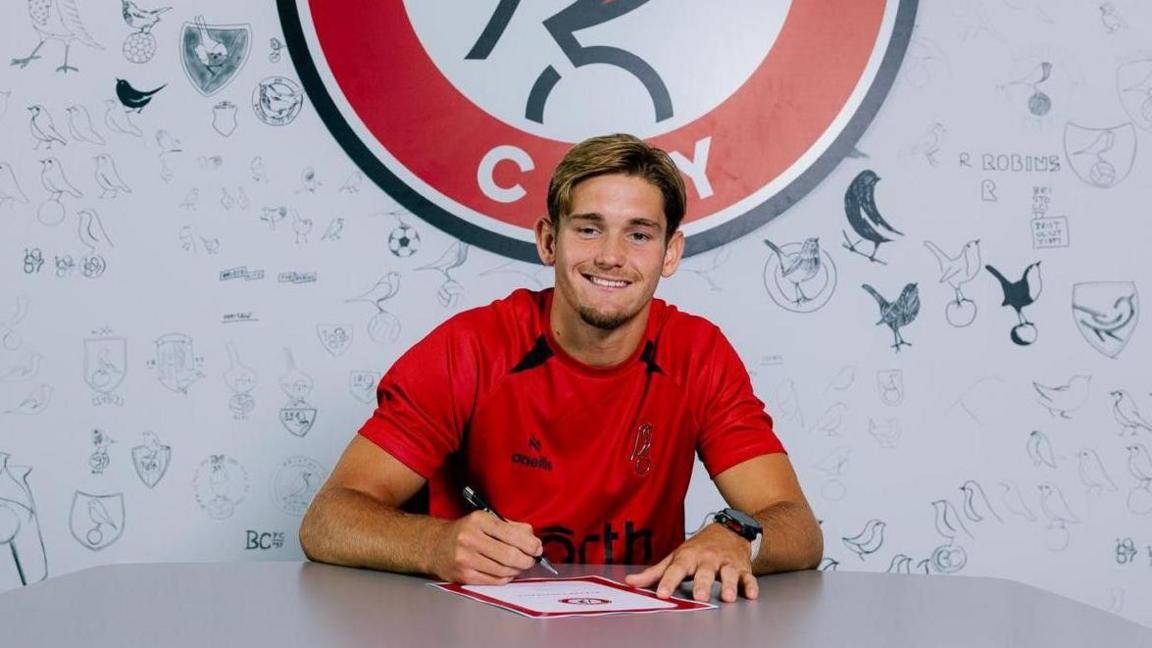 Dom Ballard sitting at a desk with a pen in his hand and a piece of paper in front of him. He is wearing a red t-shirt with a Bristol City badge on it