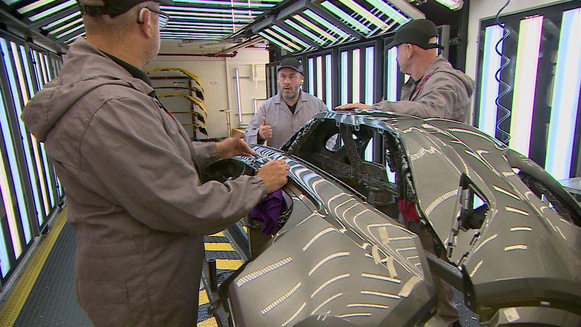 Three members of Nissan production line staff communicating using sign language. The men are all wearing grey overalls and black caps.