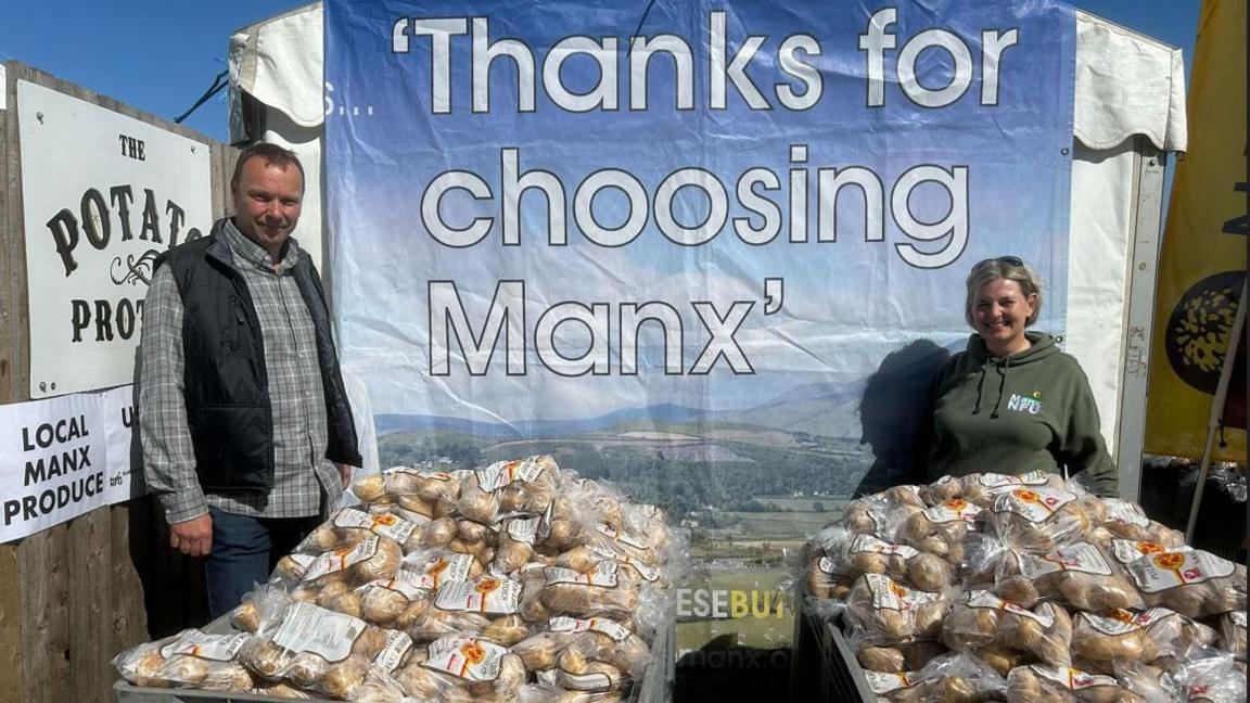 Ean Parsons and Sarah Comish stand next to large grey containers filled with clear plastic bags of potatoes. They are in front of a blue sign with white writing that reads "thanks for choosing Manx".