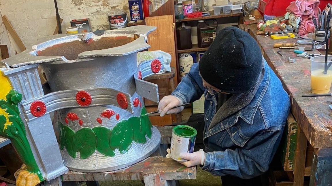 The face of the man is not visible as his head is turned towards a section of the water feature. He has a black woolly hat on and a denim jacket and is wearing white protective rubber gloves. He is holding a paint pot and brush and is dabbing on green paint.