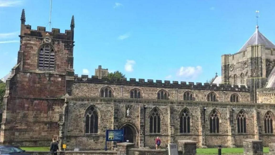 An exterior view of Bangor Cathedral, it is a clear day and there are two people in the foreground of the picture, one walking towards the front door of the cathedral. The stone cathedral has two rows of intricate arched windows and two towers at each end of the building.