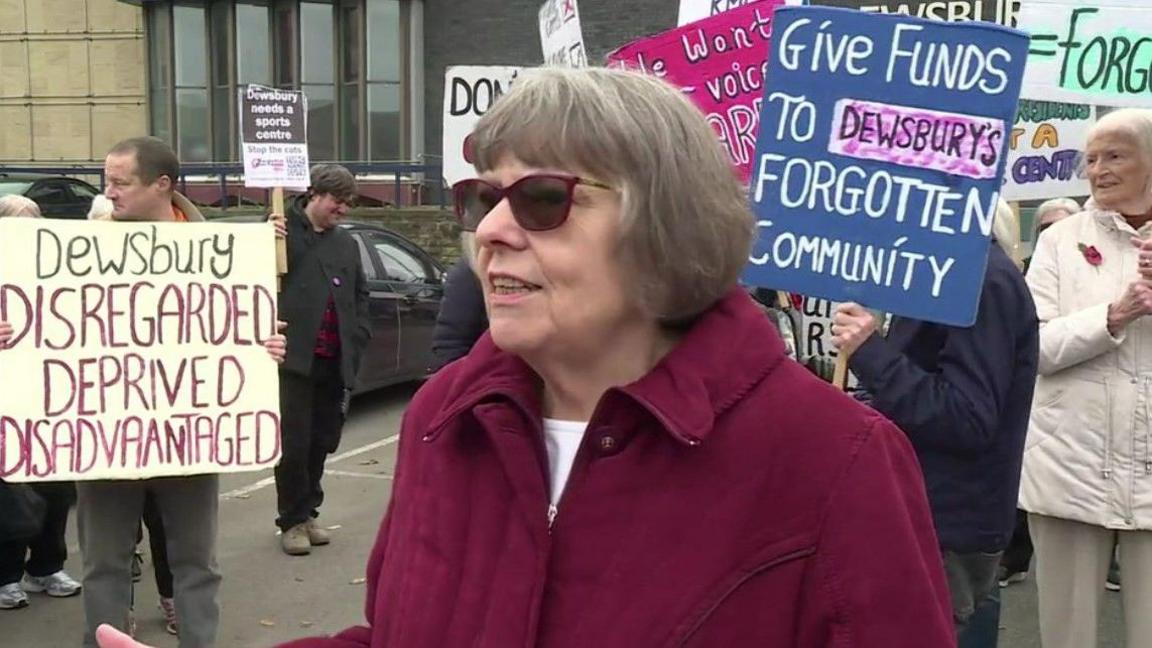 An older woman in a red coat and sunglasses with grey hair stood in front of people holding placards.