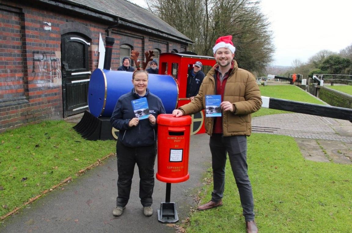 People from the Canal & River Trust stand near a stable block on the left, with a canal on the right of the photo. Two of the people are standing near the front by a postbox, with the man wearing a Santa hat.