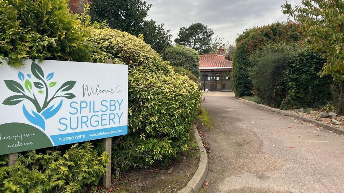 A rectangular sign sitting amongst bushes on a grass verge which reads 'Welcome to Spilsby Surgery'. A pathway is next to it leading up to a single-floor bulding at the end.