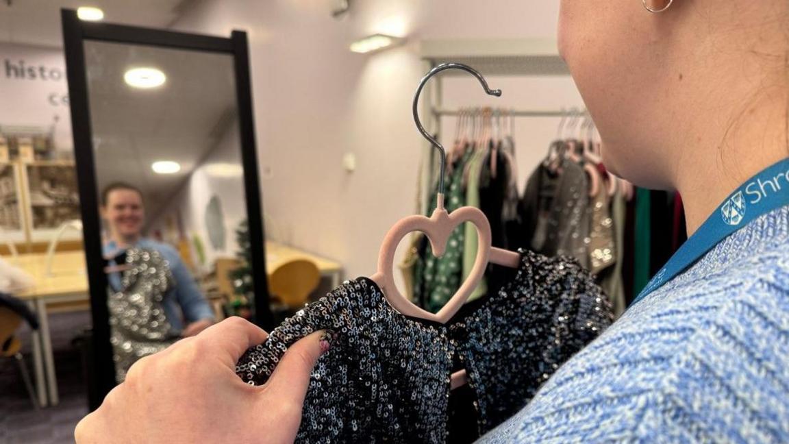 A side/back view of Siobhan Shaw holding a dress on a hanger in front of a mirror. Her face is visible in the mirror. Dresses are hung up in the background of the photo.