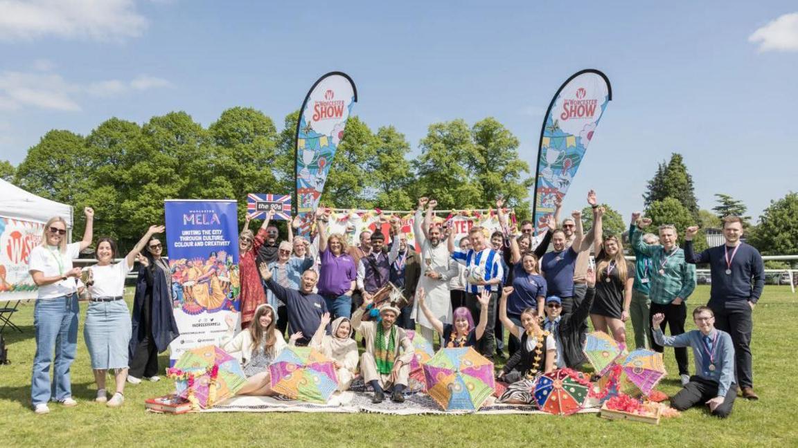 People gather at a previous Worcester Show, some are wearing costumes and some holding signs and banners that say Mela or Worcester Show. They have their arms in the air and there is a festival atmosphere, with a marquee behind, and they are looking happy. They're outside, on grass, and it is a sunny day.