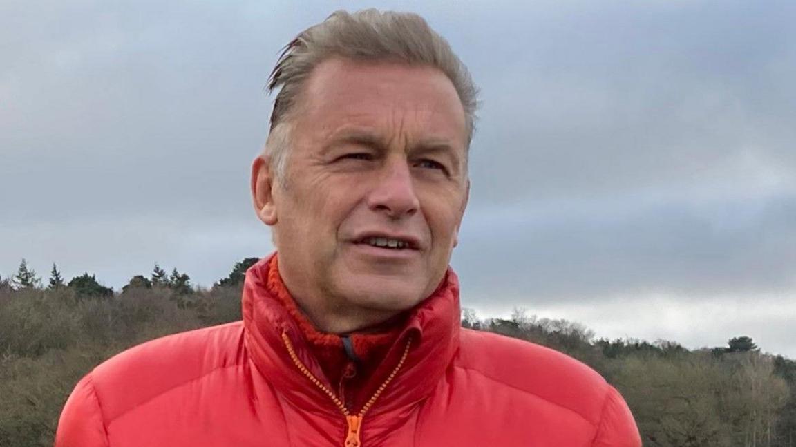 Chris Packham wearing a red padded coat. He has short grey hair and is standing outside with a forest in the distance