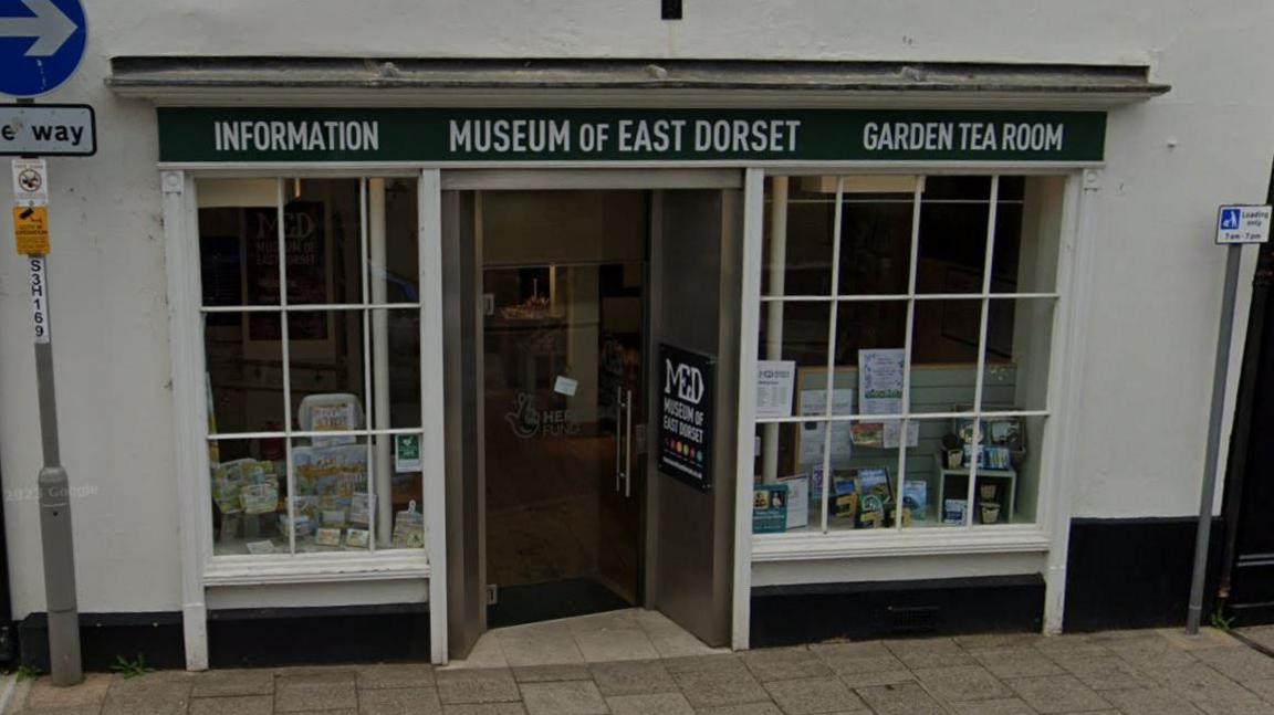 A white building with a glass shop front and a green sign above it saying "Museum of East Dorset"