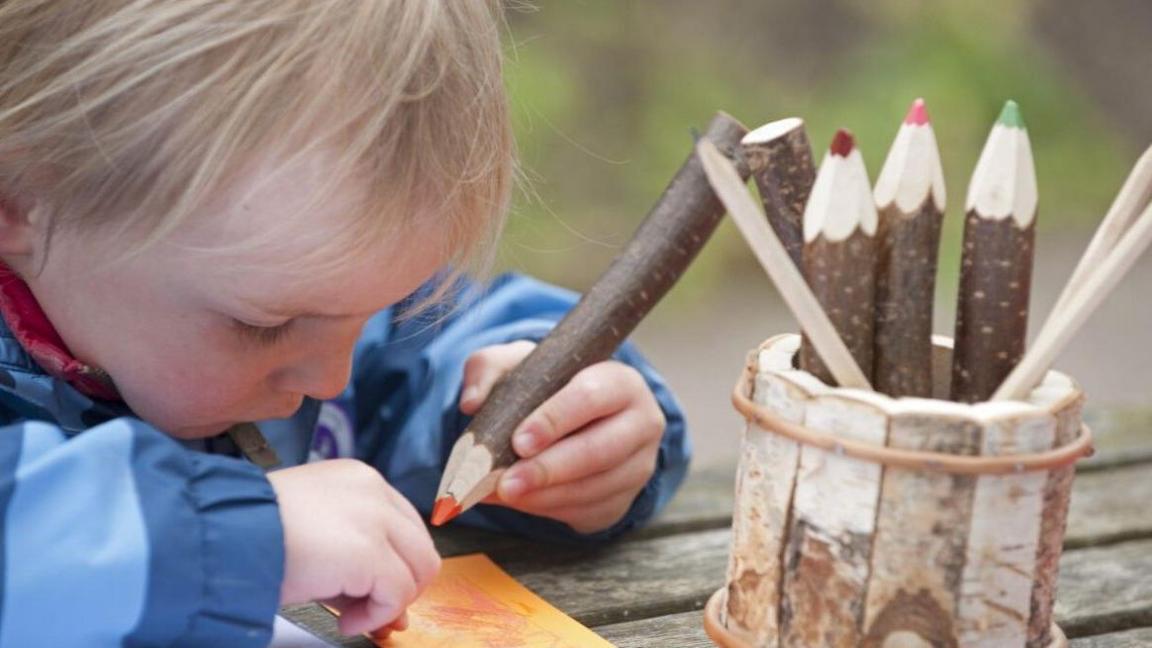 A small boy is sitting at a bench in the open air. He has fair hair and is wearing a blue anorak. He is drawing on paper using a pencil carved from a twig. Other pencils can be seen in a wooden container on the table