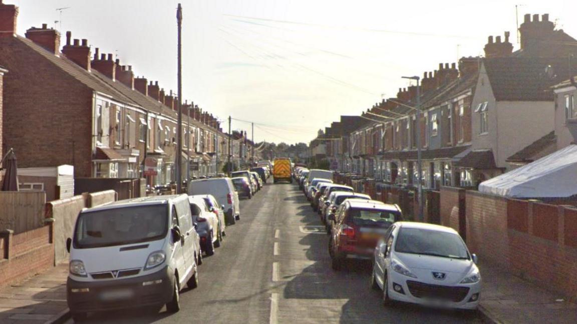 A residential street of terraced houses with cars parked in front on both sides of the road. In the far distance is a van with red and yellow emergency chevrons painted on the rear doors.