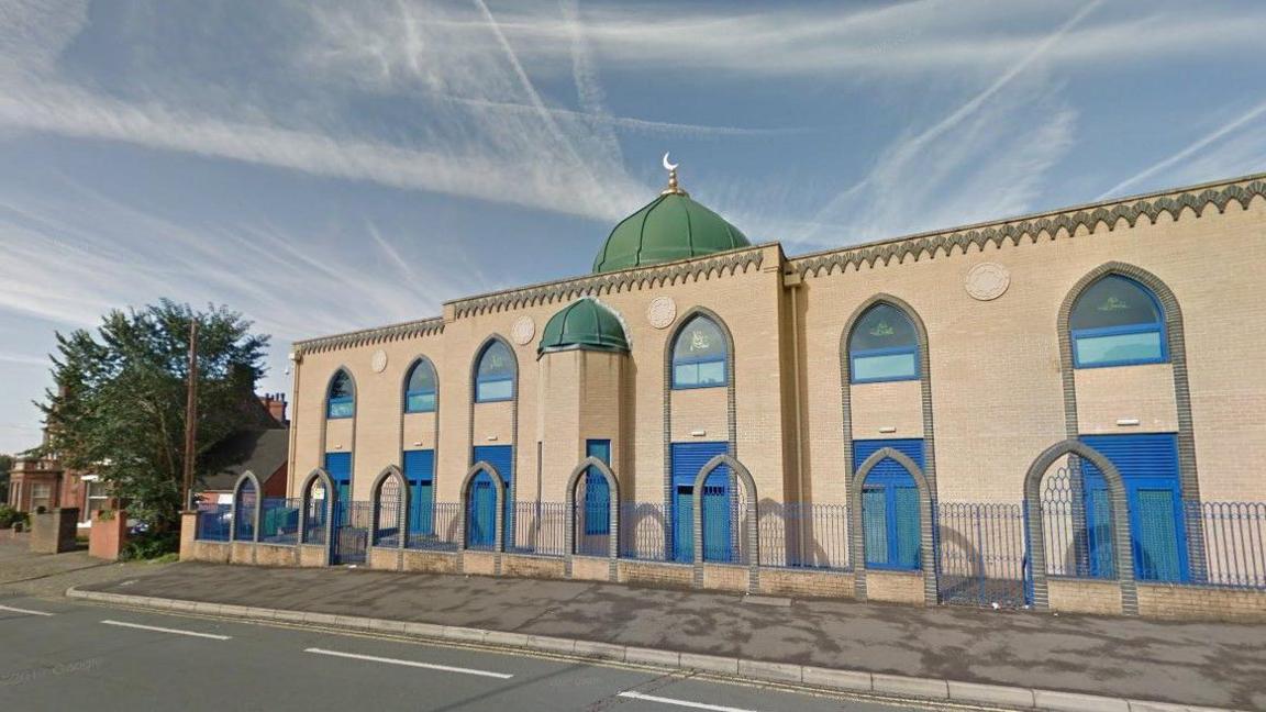 City Central Mosque, a brick-built building with green domed roof and religious symbols on rounded windows. There is a fence with grey brick arches along its length.