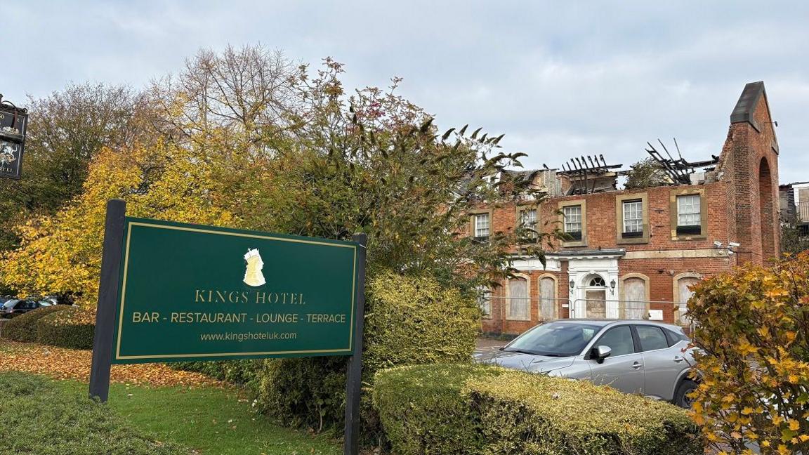 Two-storey hotel building with the upper storey badly damaged by fire. The roof has caved in. There is a green KING'S HOTEL sign in the foreground, with a tree immediately behind it. A grey car is parked in front of the hotel.