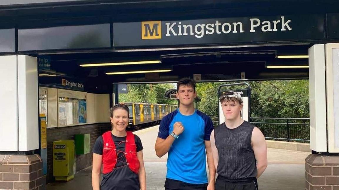 Three young people standing in front of Kingston Park Metro Station. From the left, a young woman in a red jacket, a young man in a blue top and another young man in a black vest. A metro train can be seen in the station behind them.