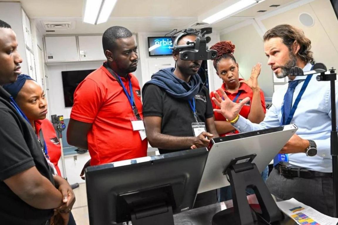 Several people gather around some monitors, while a man in a blue shirt and tie speaks to them. One wears a device on his head which has various lenses on it. They are inside a plane.