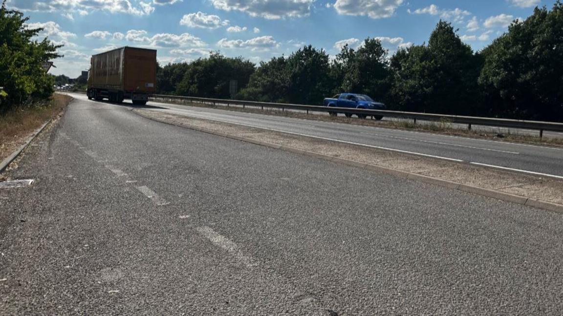Busy dual carriageway with a layby to the left of the image. There is a lorry driving away from the camera and a car is on the other lane