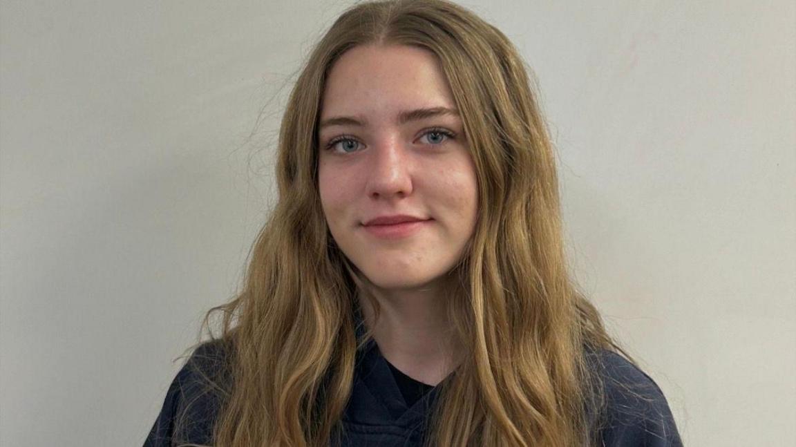 An image showing Keegan Radomski. She has long slightly wavy blonde hair, blue eyes and is wearing a black shirt. Behind her is a plain, white backdrop. 