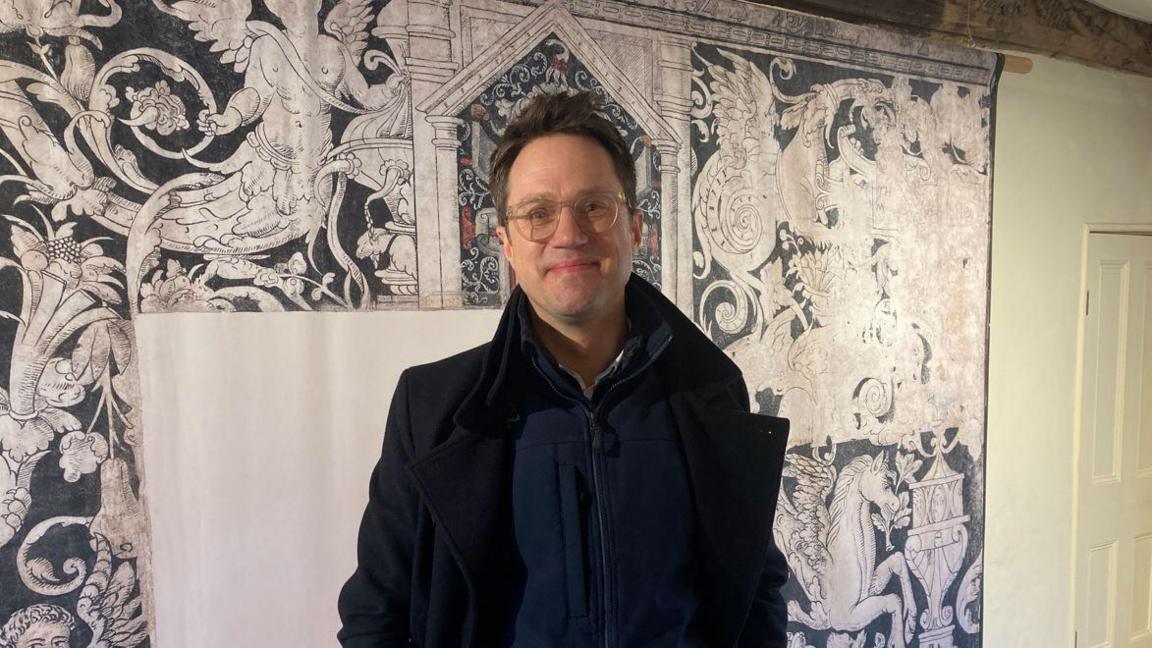 A man with dark hair, glasses and a dark coat standing in front of a decorative wall painting