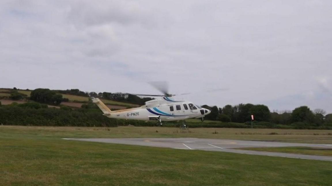 A helicopter hovering above a tarmac square in a green field, with trees behind.