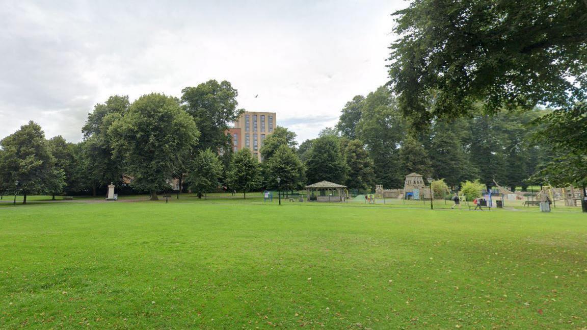 A Google Street View of a city park. Trees are along the edge of the park and a multi-storey building is visible beyond. A large grass area is in the foreground, and a playground with wooden structures is behind it.