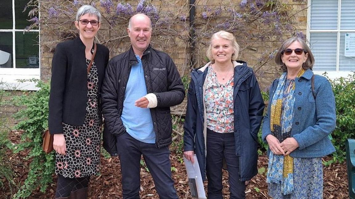 There are four people standing in a line in front of a building that has purple flowers on the walls and greenery. Left to right, the people are Joanne Willett, Philip Wylie, Alison Barr and Sheila White. They are all looking at the camera and smiling.