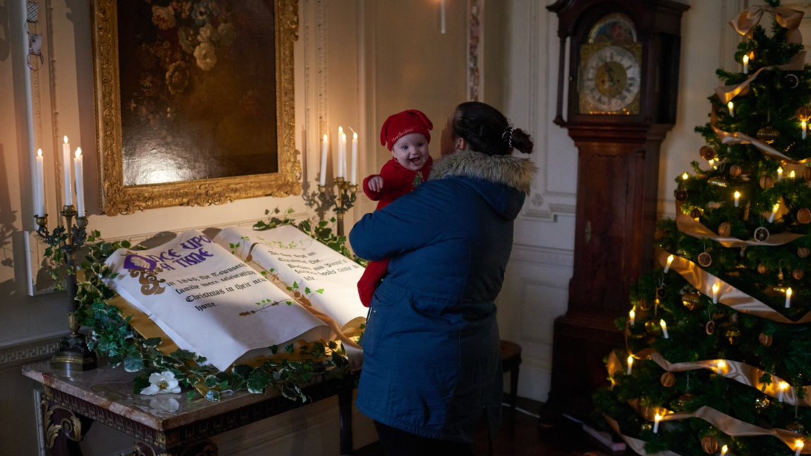 One of the rooms in Mompesson House with a mother holding a baby and looking at an oversized book of fairytales laid out on a table with candles and a Christmas tree and decorations in the background