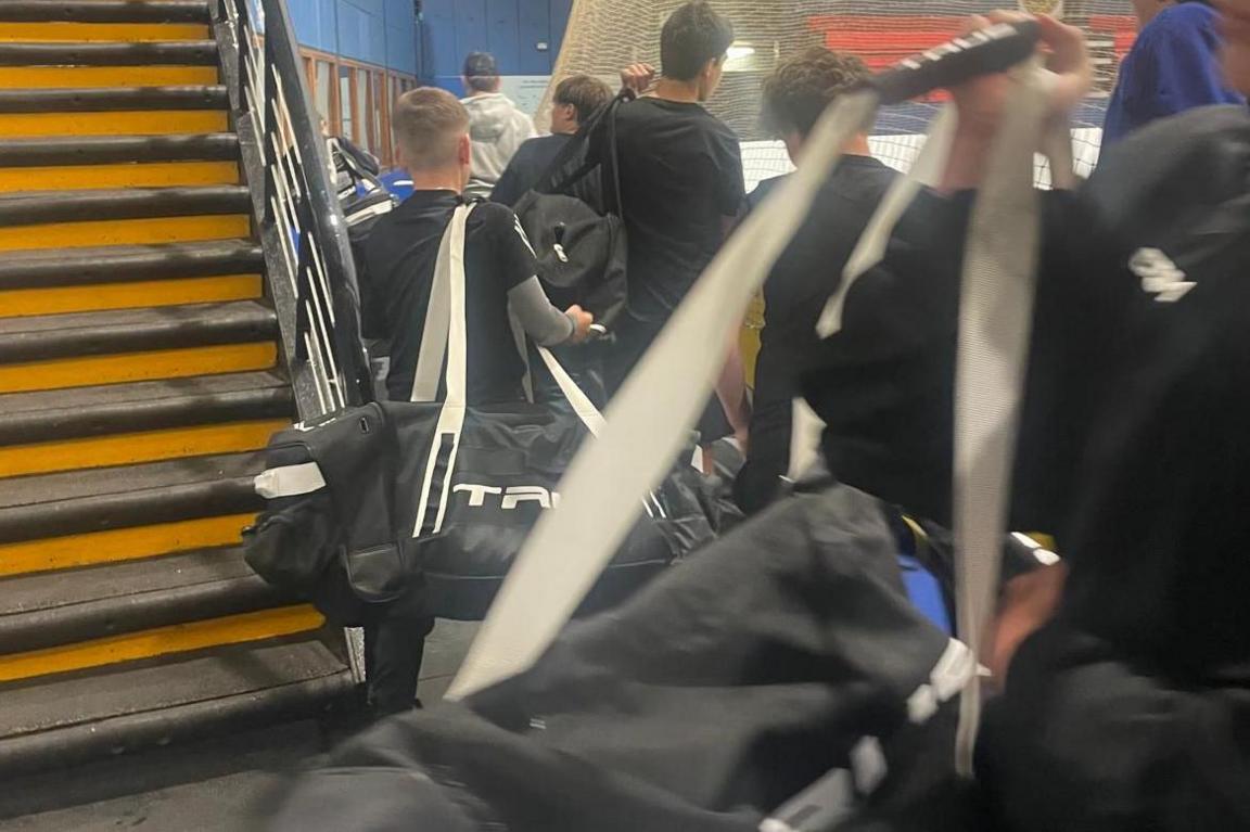 Boys approach the ice with large kit bags over their shoulders.