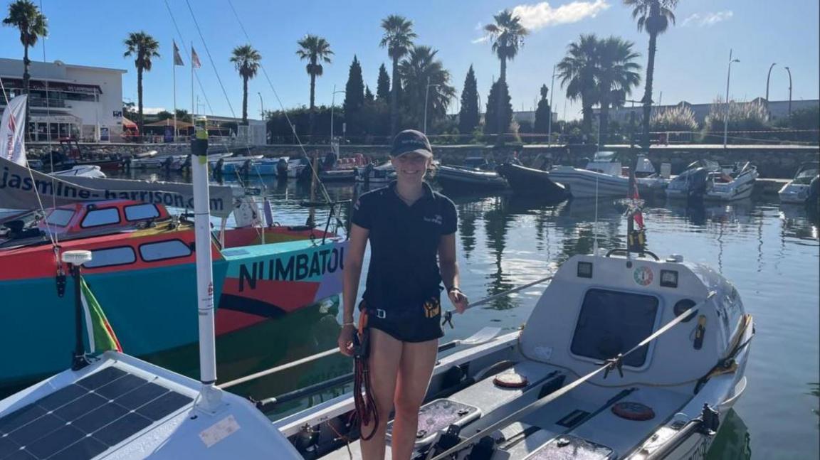 Zara Lachlan wearing her hair tucked into a dark blue baseball cap and a dark blue polo shirt, both with Team Forces and the Union flag logos. She is standing in a white boat just before she set off on the challenge. It is in a marina with other boats around it.