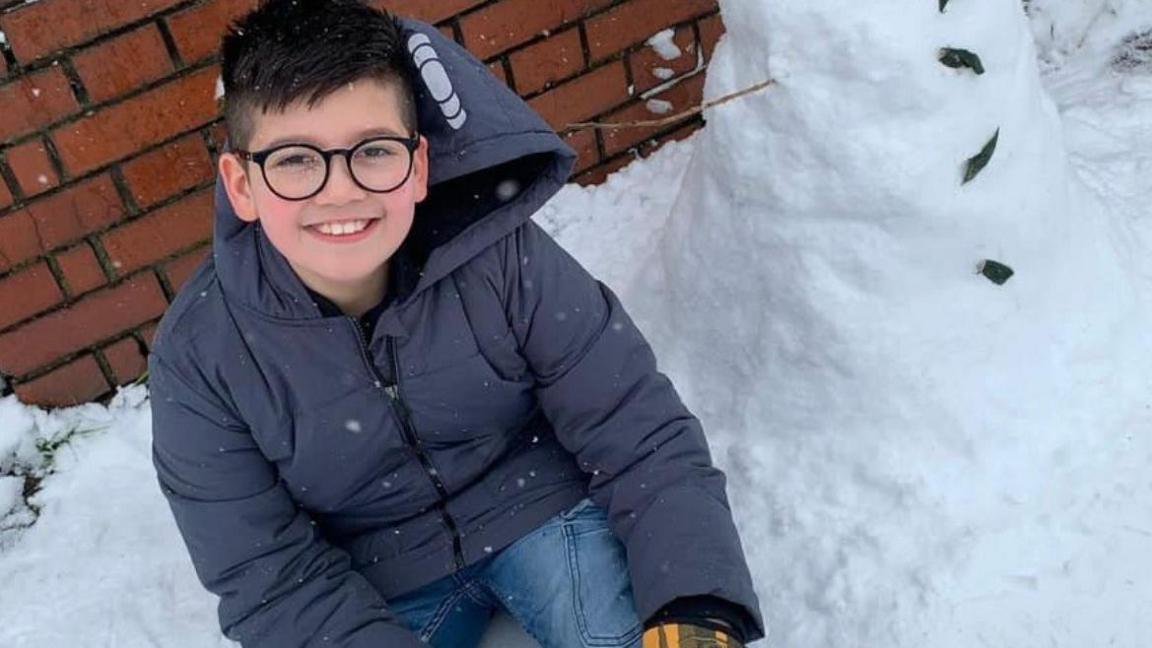 Twelve-year-old Azaan Khan sits next to a snowman.