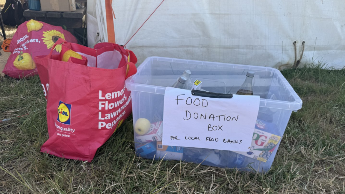 A clear plastic box with a handwritten paper sign attached to it saying "Food donation box for local food banks" on the grass outside a tent in a campsite at Glastonbury. It contains some bottles of soft drink, some tinned food and a box of tea bags. There is a red Lidl Bag for Life next to it.