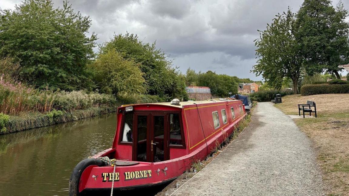 A red narrowboat with the name "The Hornet" written on it is tied up next to a gravel towpath on a canal